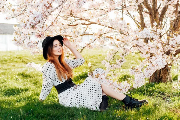 Jonge brunette vrouw met hoed en jurk zitten in de buurt van witte roze bloeiende boom. Generatie Z meisje geniet van de voorjaarsstemming tijdens een zonnige dag. Ontspan je op de natuur. Voorjaarsbloesem. Selectieve focus. — Stockfoto
