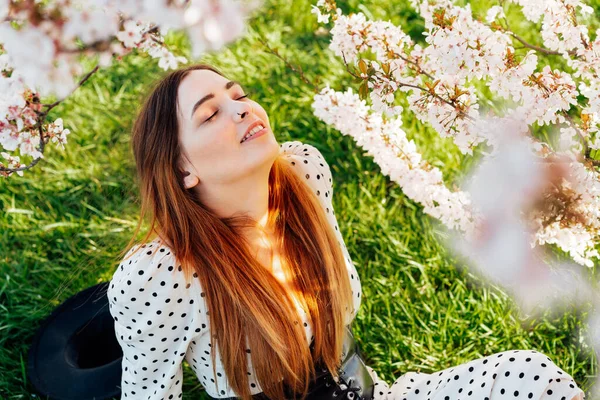 Jovem morena de vestido com os olhos fechados sentados perto de uma árvore rosa florescendo e desfrutando do sol. Geração Z menina desfrutar de humor primavera durante um dia ensolarado. Relaxe na natureza. — Fotografia de Stock