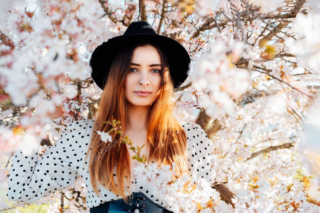 Young brunette woman in hat and dress in white pink blooming tree. Generation Z girl enjoy spring mood during sunny day. Springtime blossom. Selective focus, copy space