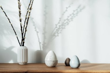 Home interior with easter decor. Pussy willow branches in a vase, easter eggs statuettes on the wooden console with light and shadow background. Minimalism, scandinavian style. selective focus.