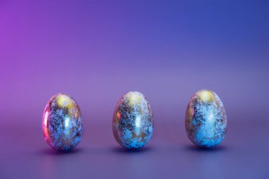Farklı desenlere sahip üç Paskalya altın yumurtası, neon renkli ışık. Mor, minimalist renk arkaplanı. Modern Paskalya modası. Seçici odaklanma. Boşluğu kopyala.