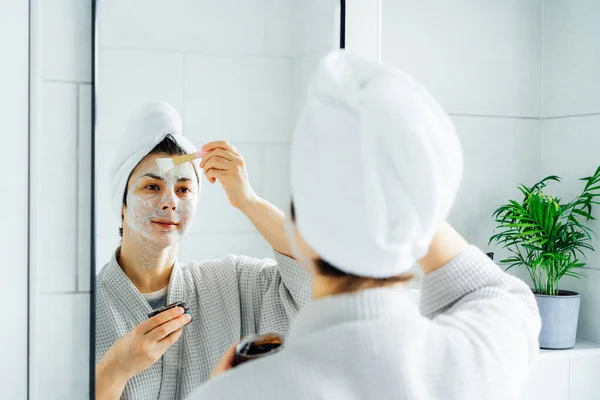 Mujer en albornoz con una toalla en la cabeza mirando en el espejo y la aplicación de mascarilla de arcilla cosmética facial en su cara en el baño. El concepto de procedimientos cosméticos para el cuidado de la piel. Home Belleza autocuidado — Foto de Stock