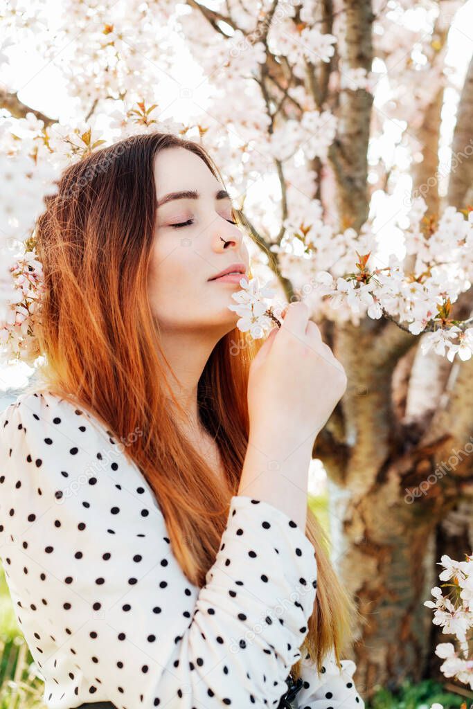 Young brunette woman with closed eyes sitting near a pink blooming tree and enjoying the moment. Generation Z girl enjoy spring mood during a sunny day. Relax, freedom and personal fulfillment.