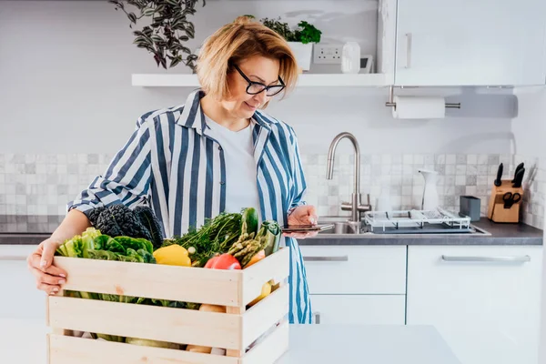 Livraison de nourriture à domicile en ligne. Femme vérifiant sa liste de commande en ligne sur son téléphone. Boîte en bois avec légumes frais et fruits debout sur la table de cuisine. Nourriture locale. Nouveau départ d'une vie saine. — Photo