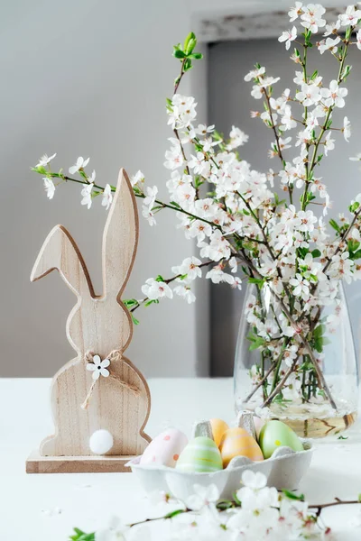Vazosunda çiçekler olan ağaç dallı ahşap Paskalya tavşanı ve beyaz masadaki renkli paskalya yumurtaları. Mutlu Paskalya bahar dikey kartları. Seçici odaklanma. Boşluğu kopyala. — Stok fotoğraf