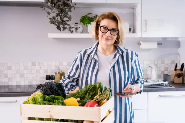 Livraison de nourriture à domicile en ligne. Femme vérifiant sa liste de commande en ligne sur son téléphone. Boîte en bois avec légumes frais et fruits debout sur la table de cuisine. Nourriture locale. Nouveau départ d'une vie saine — Photo