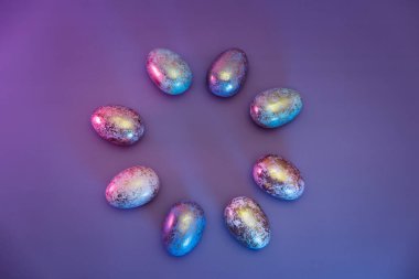 Üst çerçeve, Paskalya altın yumurtalarından yapılmış. Farklı desenlerde, neon renkli ışık. Mor, minimalist renk arkaplanı. Modern Paskalya modası. Seçici odaklanma. Boşluğu kopyala.