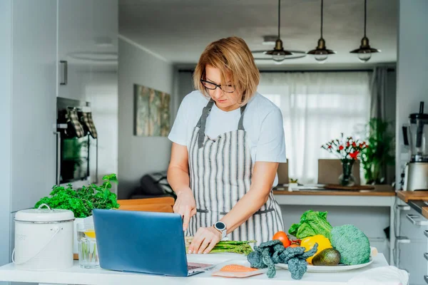 Femme d'âge moyen suivant un cours vidéo tutoriel de cuisine sur ordinateur portable tout en préparant le repas dans une cuisine. Femme cuisine plat sain, poisson et légumes sur la table. Recette en ligne. Concentration sélective. — Photo