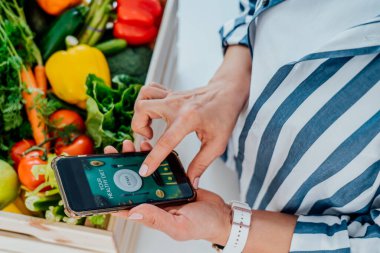 Üst görüşlü kadın elinde taze sebzeli ahşap kutu ve sağlıklı beslenme programı ile aktif online mobil uygulamalı telefon tutuyor. Sağlıklı bir yaşam tarzının yeni başlangıcı, kilo verme kavramı