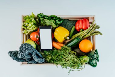 Üst manzara ahşap kutu, taze sebzeler ve beyaz arka planda beyaz ekranlı telefon. Diyet programı, yemek teslimatı, kilo kaybı için çevrimiçi mobil uygulama modeli. Sıfır atık