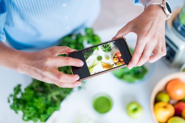 Sosyal medya için içerik oluşturan blogcu. Kadın fotoğraf çekiyor, video çekiyor. Mutfak masasında bir bardak detoks yeşili smoothie ve malzemeleri var. Sağlıklı beslenme, yemek pişirme, kilo verme günlüğü.