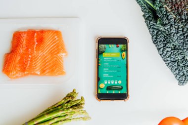 Keto yemek diyeti planlaması. Mobil uygulamalı keto menüsü olan Smartphone taze malzemeler, taze sebzeler ve somon balığı ile birlikte masada. Pişirmeye hazır. Sağlıklı yaşam tarzı, kilo kaybı. Tarif kutusu.