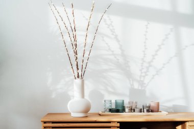 Modern minimalist Scandinavian style interior. Blooming branches of the pussy willow in ceramic vase and tray with candles on a wooden console with sunlight and shadows on white gray wall. Copy space.
