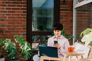 Bilgisayarlı bir kadın telefonda sosyal ağları kaydırıyor, sohbet ediyor, tatilde evde çay içerken ve dinlenirken video izliyor. Ev bitkileriyle dolu rahat, modern çatı katı tarzı bir oda. uzak işyeri