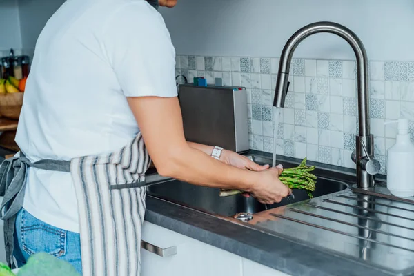 Woman washing fresh asparagus bunch under running water over sink on her kitchen. Preparing food ingredients for lunch. Cooking healthy food. Recipes with vegetables, balanced diet. — Stock Photo, Image