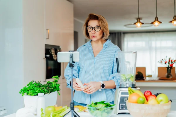 Les femmes du Moyen Âge blogue pour son canal de style de vie de bien-être sur la vie saine et équilibrée dans la cuisine de sa maison. Création de contenu vidéo pour les médias sociaux avec caméra téléphonique sur un trépied. — Photo