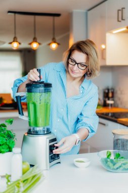 Evde blender ve yeşil sebzeli orta yaşlı bir kadın detoks veya yeşil smoothie yapıyor. Sağlıklı beslenme, yemek, yemek. Doğal anti-yaşlanma, kilo verme programı. Vejetaryen diyeti. Dikey kart.