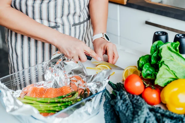 Baked salmon with green asparagus recipe steps. Step four. Woman slice fresh lemon to add to salmon and veggies in the baking form. Step by step recipe. Healthy cooking at home according to recipe — Stock Photo, Image