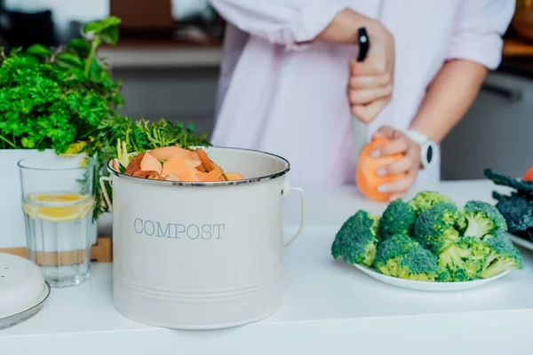 Compost the kitchen waste, recycling at home. Compost bin with vegetables cutted leftovers on the kitchen table with blurred woman cooking meal. Environmentally responsible, ecology. Selective focus. — Stock Photo, Image