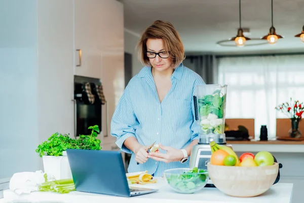 Femme d'âge moyen regarder un cours vidéo tutoriel de cuisine sur ordinateur portable tout en préparant un repas dans une cuisine. Une femme qui fait un smoothie vert. Recette en ligne. Mode de vie sain, programme de perte de poids. — Photo