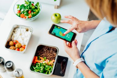 Kilo vermek için sağlıklı bir diyet planı, günlük hazır yemek menüsü. Yemek takip uygulaması kullanan bir kadın mutfak tartısı için önceden pişirilmiş yemek kutusunu tartarken. Tabakla dengeli porsiyon. Pişirme öncesi