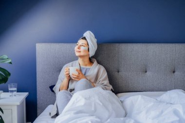 Pijamalı, bornozlu, göz bantlı ve kafasında havluyla sabah kahvesini içip dinlenen bir kadın. Sabah alışkanlıkları ve ev güzelliği rutini. Kişisel bakım. Seçici odak
