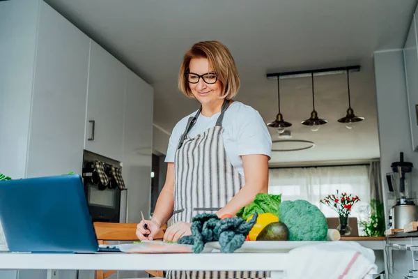 Middle age woman following a cooking tutorial video course on laptop while preparing meal in a kitchen. Woman cooking healthy dish, fish and vegetables on the table. Online recipe. Selective focus — Stock Photo, Image