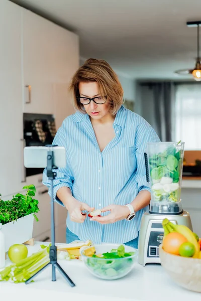 Orta yaşlı kadınlar sağlıklı yaşam tarzı kanalı için evlerinin mutfağında sağlıklı dengeli yaşam hakkında blog yazıyorlar. Üç ayaklı bir telefon kamerasıyla sosyal medya için video içeriği oluşturuluyor