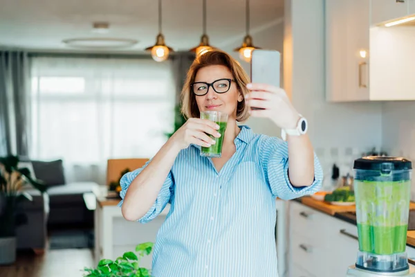 Middle aged woman making selfie with just made glass of detox shake, green smoothie in the kitchen. Healthy dieting, eating, cooking, weight loss program. Blogger creating content for social media. — Stock Photo, Image