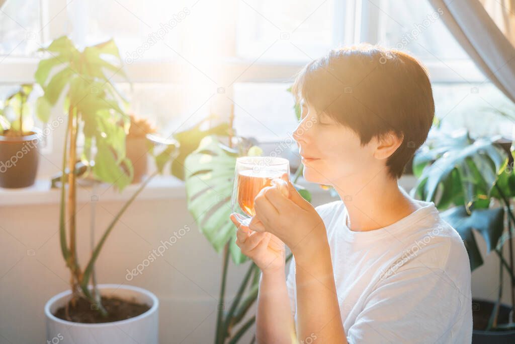 Woman with closed eyes drinking tea in a room with green house plants, enjoying its taste and aroma in a sunset or sunrise light. Relaxing and self-care, personal fulfillment. Selective focus.