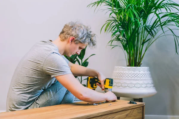 Elektrikli tornavidayla genç bir adam yeni evindeki talimatlara göre televizyon standı konsolu kuruyor. Adam evde kablosuz tornavida kullanarak mobilya monte ediyor. Seçici odak.