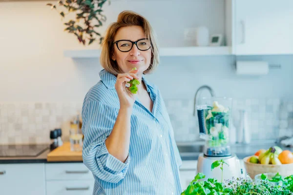 Healthy detox vegan diet. Portrait of middle aged woman eating a stick of green celery while making smoothie on kitchen. Healthy dieting lifestyle, weight loss program. Selective focus — Photo