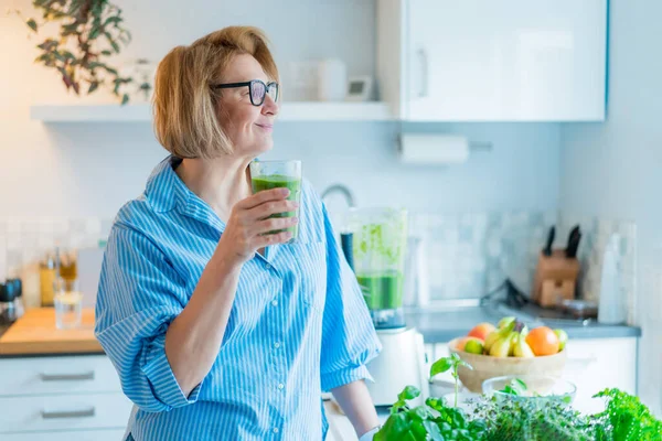 Smiling middle aged woman with just made glass of detox shake, green smoothie in the kitchen. Healthy dieting, eating, cooking. Natural anti aging methods, weight loss program. Vegan, vegetarian diet — 스톡 사진