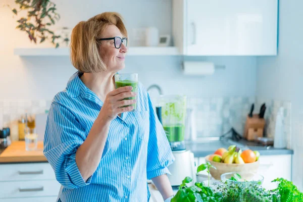 Femme d'âge moyen souriante avec un verre de désintoxication, smoothie vert dans la cuisine. Régime alimentaire sain, manger, cuisiner. Méthodes naturelles anti-âge, programme de perte de poids. Vegan, régime végétarien — Photo