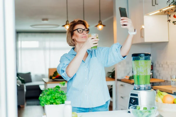 Femme d'âge moyen faisant selfie avec verre juste fait de désintoxication shake, smoothie vert dans la cuisine. Régime alimentaire sain, manger, cuisiner, programme de perte de poids. Blogueur créant du contenu pour les médias sociaux. — Photo