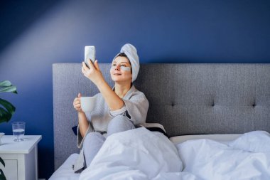 Pijamalı, bornozlu, göz bantlı ve kafasında havluyla sabah kahvesini içip yatağında telefonla selfie çeken bir kadın. Sabah alışkanlıkları ve ev güzelliği rutini. İçerik oluşturma.