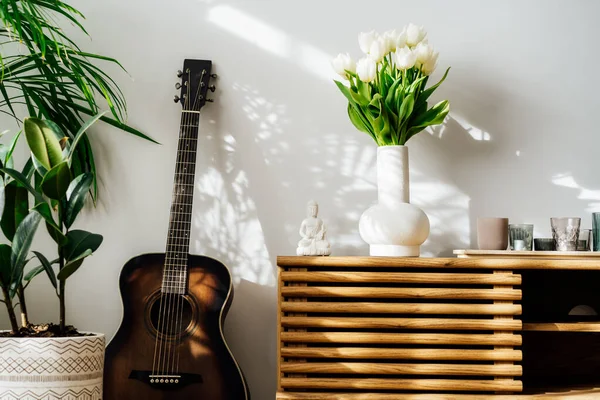 Scandinavian Minimalist home interior details - bouquet of white tulip flowers in vase, candles, Buddha statuette on wooden cabinet. Green house plants and acoustic guitar near white wall with shadow