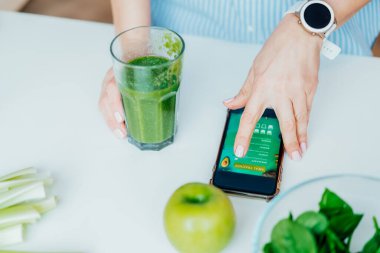 Close up woman with green smoothie fills, making notes in the meal treaker on her phone on the kitchen table. Healthy diet and balanced eating, weight loss online app program. Selective focus