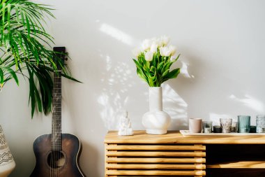 İskandinav Minimalist ev dekorasyonu ayrıntıları - vazoda beyaz lale çiçekleri, mumlar, ahşap dolapta Buda heykelciği. Yeşil ev bitkileri ve akustik gitar, gölgeli beyaz duvarın yanında..