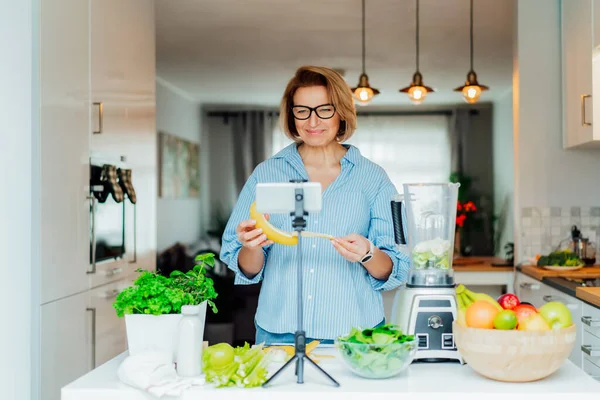 Les femmes du Moyen Âge blogue pour son canal de style de vie de bien-être sur la vie saine et équilibrée dans la cuisine de sa maison. Création de contenu vidéo pour les médias sociaux avec caméra téléphonique sur un trépied — Photo