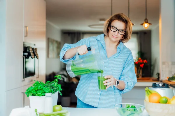 Middle aged woman pouring detox drink, green smoothie from blender into glass at the kitchen. Healthy dieting, eating, cooking. Natural anti aging methods, weight loss program. Vegan, vegetarian diet — 스톡 사진