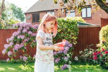 Bahçedeki yeşil bir çimenlikte oynarken baloncuklar çıkaran tonlu kızıl saçlı tatlı küçük kız. Okul tatilinde evde eğleniyorum. Çocuklar için çevrimdışı aktivite. Mutlu çocukluk kavramı
