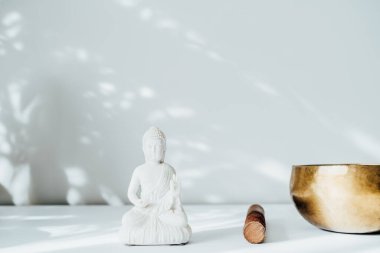 Dekoratif beyaz Buda heykelciği ve beyaz arka planda güneş ışığı gölgeli Tibet şarkı kasesi. Meditasyon ve rahatlama ayini. Egzotik masaj. Minimalizm. Uzayı kopyala Seçici odak