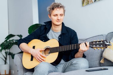Adamın biri telefonuyla internetten gitar dersi alıyor. Gri koltukta oturuyor. Oturma odasındaki kanepe. Akustik gitar dersi izliyor. Yeni hobi. Seçici odak.