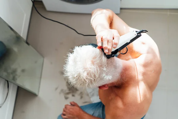 Top view λευκό, ασημένια μαλλιά άνθρωπος κάνει αυτο-κούρεμα με ένα clipper και κοιτάζει στον καθρέφτη. αντρική αυτοφροντίδα στο σπίτι με ηλεκτρικό ξυράφι. Επιλεκτική εστίαση. — Φωτογραφία Αρχείου