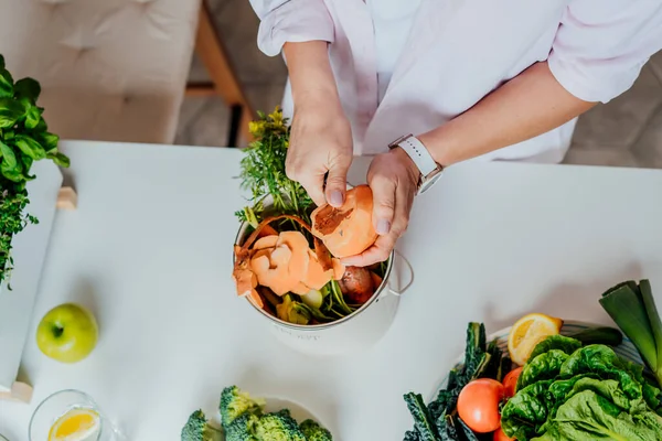 Compost the kitchen waste, recycling at home. Top view Woman cleans sweet potato and puts vegetables cutted leftovers into the garbage, compost bin on her kitchen. Environmentally responsible, ecology