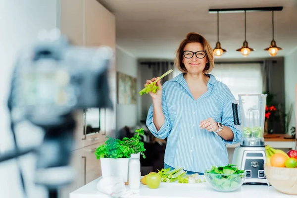 Femme d'âge moyen blogue pour son canal de style de vie de bien-être sur la vie saine et équilibrée dans la cuisine de sa maison. Création de contenu vidéo pour les médias sociaux avec caméra sur un trépied — Photo