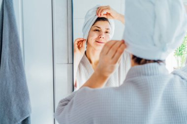 Sakin, gülümseyen, spa bornozu ve havluyla evde duş aldıktan sonra rahatlayan melez bir kadın. Aynadaki yansıma. Güzellik tedavisi konsepti. Vücut derisi ve saç bakımı.