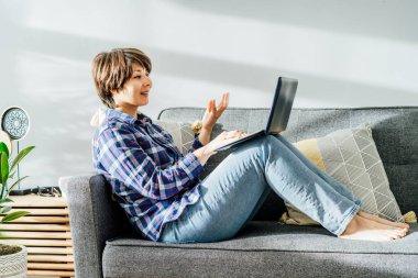 Duygusal bir kadın laptopta video görüşmesi yapıyor. Yumuşak koltukta oturuyor. Modern, yeşil bitkilerle dolu bir iç mekanda. Uzaktan kumanda. Evdeki rahat çalışma alanı. Çevrimiçi iletişim, arkadaşlarla sanal buluşma.