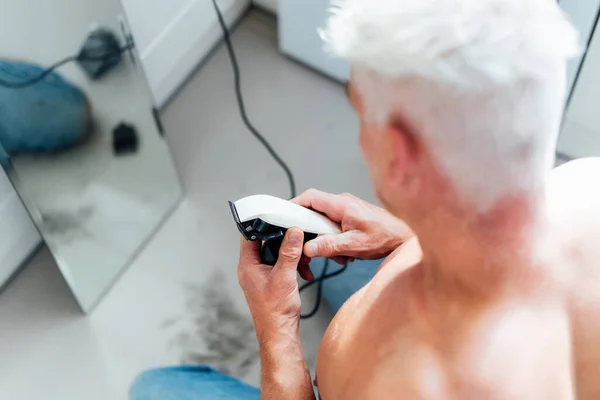 Vista posterior blanco, hombre de pelo plateado haciendo auto corte de pelo con un clipper y se mira en el espejo. autocuidado masculino en casa con afeitadora eléctrica. Enfoque selectivo. — Foto de Stock